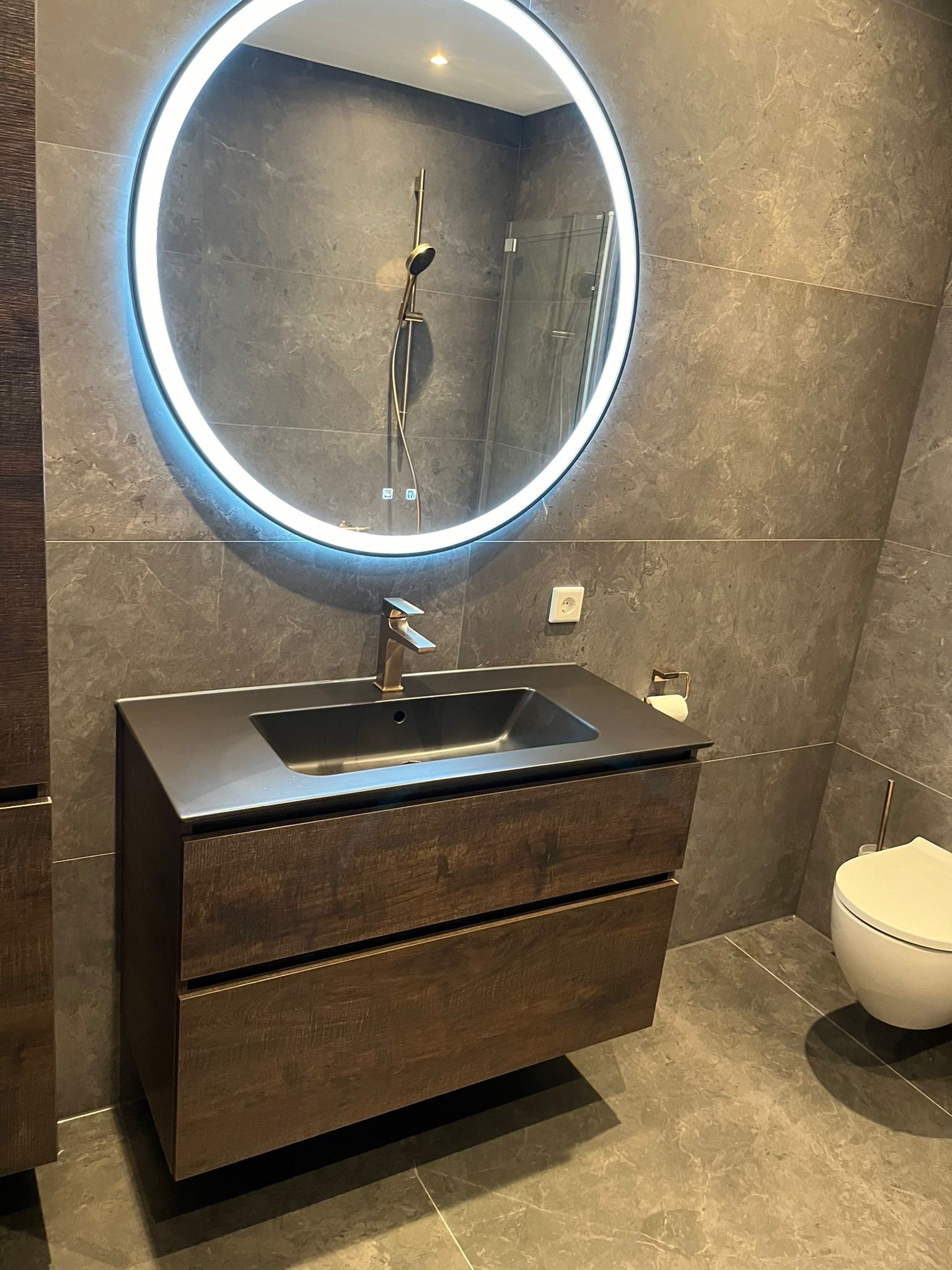 Badkamer met donkergrijze marmerlook tegels, een ronde verlichte spiegel en een donker houten badmeubel